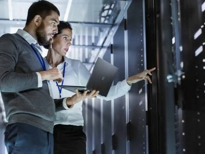 Male IT specialist holding a laptop discussing server support, virtual machines, and data centers with a female colleague. Managed IT Services